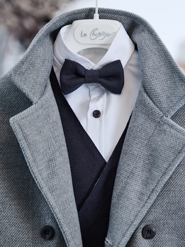 Μια κλασσική πρόταση για βάπτιση, αποτελούμενη από έξι τεμάχια. Το λευκό βαμβακερό πουκάμισο αποτελεί την τέλεια βάση για ένα υπέροχο σταυρωτό γιλέκο, από navy blue πικέ ύφασμα. Ένα βελούρ διαγωνάλ παλτό, τύπου μοντγκόμερι σε γκρι αποχρώση, σετάρετε υπέροχα με το άνετο παντελόνι, από μαλακό πικέ ύφασμα. Το σύνολο ολοκληρώνεται από αξεσουάρ όπως παπιγιόν και τιράντες που δίνουν έναν ναυτικό αέρα στο σύνολο.