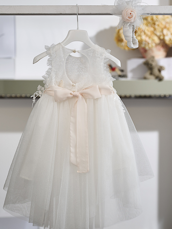 Η διαχρονική κομψότητα και η εκλεπτυσμένη αισθητική χαρακτηρίζουν αυτό το εντυπωσιακό βαπτιστικό φόρεμα σε off white απόχρωση. Το κορσάζ είναι διακοσμημένο με εκλεπτυσμένη δαντέλα και ανάγλυφα απλικέ μοτίβα, που χαρίζουν πλούσια υφή και ιδιαίτερο χαρακτήρα. Στη μέση, μια σατέν ζώνη σε απαλό σομόν τόνο στολίζεται με χειροποίητα λουλούδια και διακριτικές λεπτομέρειες από τούλι, προσθέτοντας μια κομψή χρωματική πινελιά.